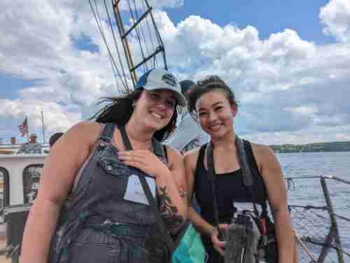 Two students smile for the camera. They are on a boat with masts. There are puffy white clouds and a blue sky. 
